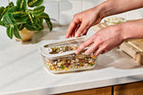 Stack of Naked Kitchen glass food containers with food inside on a refrigerator shelf.