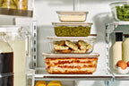 Stack of Naked Kitchen glass food containers with food inside on a refrigerator shelf.
